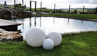 Three decorative spheres are placed on a grassy lawn next to a pond. The pond has a wooden deck and a small waterfall.