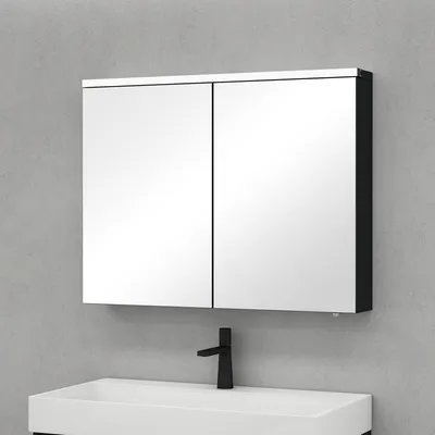 A mirrored medicine cabinet with two doors is mounted on a light gray wall above a white sink with a black faucet.