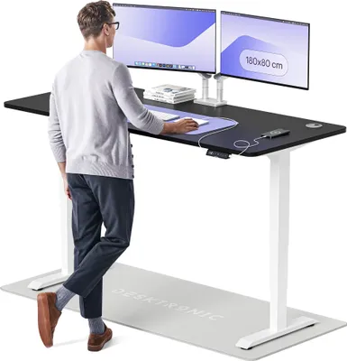 A man stands at a height-adjustable desk with two monitors, using a mouse and keyboard. A charging mat is on the desk, and a standing mat is on the floor.