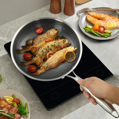 A hand is holding a frying pan with three cooked fish, tomatoes, and lemon slices, placed on a black induction cooktop. Another plate with cooked salmon and asparagus is visible in the background.