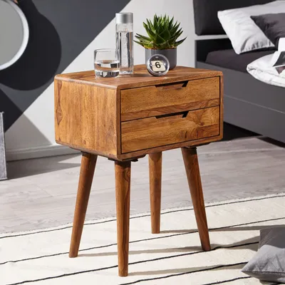 A wooden nightstand with two drawers is placed next to a bed, decorated with a plant, a glass of water, and a bottle.