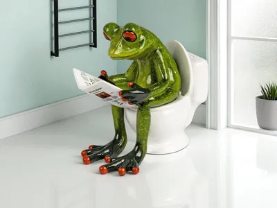 A green frog figurine sits on a toilet and reads a newspaper.