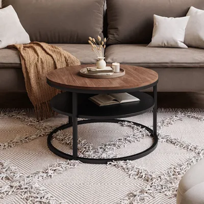 A round coffee table with a wooden top and black metal frame is placed on a patterned rug in front of a sofa.