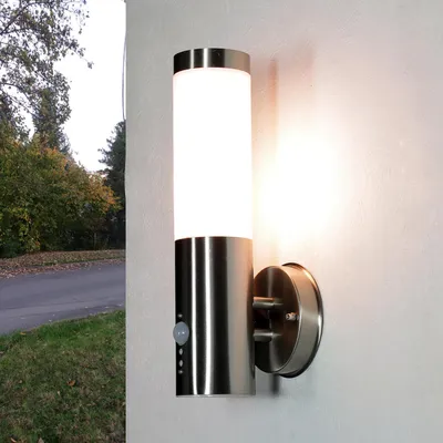 A modern stainless steel outdoor wall light with a motion sensor is mounted on a textured wall. The light is illuminated, casting a warm glow. To the left, a road and trees with autumn foliage are visible.