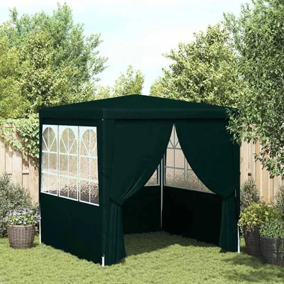 A dark green party tent is set up on a grassy lawn in a backyard, surrounded by trees and a wooden fence.