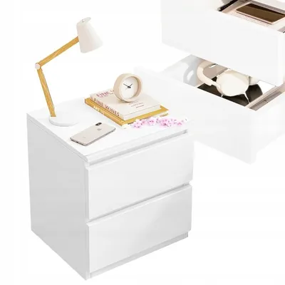 A white bedside table with two drawers, a lamp, books, a clock, and a smartphone on top.
