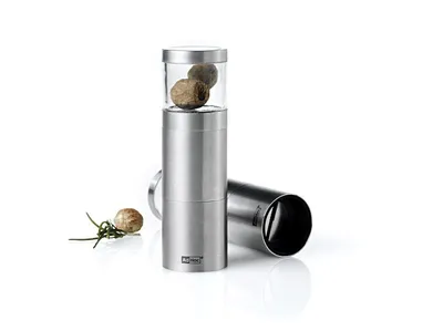 A stainless steel nutmeg grater with a glass top filled with nutmegs, placed on a white background with a sprig of rosemary and a single nutmeg to the side. Another part of the grater is lying on its side in the background.
