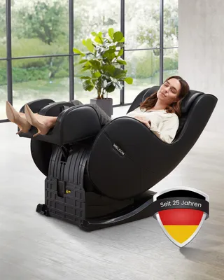 A woman is relaxing in a black massage chair in a well-lit room with large windows overlooking a garden.