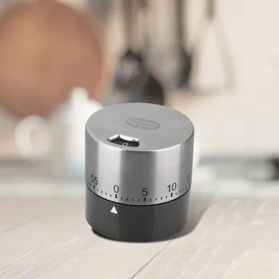 A mechanical kitchen timer sits on a wooden surface with a blurred background of kitchen items.
