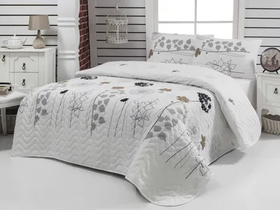 A neatly made bed with a white quilted bedspread featuring a floral pattern in gray, black, and tan. The bed is set in a bedroom with white paneled walls and a bedside table.