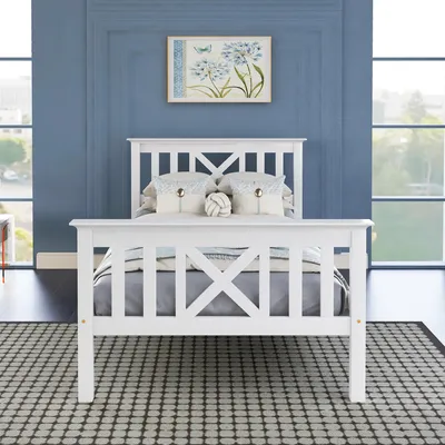 A white wooden bed frame with a slatted headboard and footboard, set against a blue wall with a framed picture of flowers above it. A patterned rug is on the floor in front of the bed. Large windows are on either side of the bed.