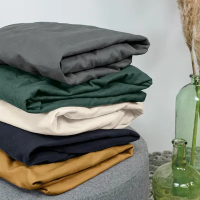 A stack of folded bedding in various colors, with a decorative vase and dried grass in the background.