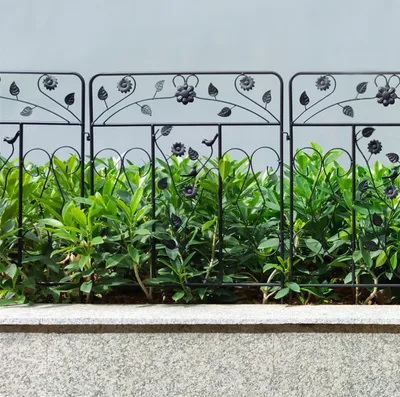 A black metal garden fence with floral and bird motifs, separating a bed of green plants from a stone wall.