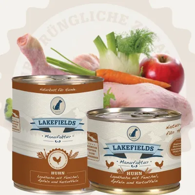Two cans of dog food are displayed in front of raw ingredients like chicken, carrots, fennel, and an apple.