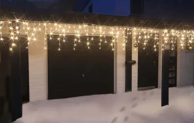 Icicle lights are hung on the eaves of a house above a garage and two doors, with snow on the ground.