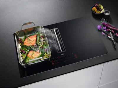 A modern kitchen countertop with a black induction cooktop featuring an integrated downdraft ventilation system. A metal roasting pan with salmon and vegetables is placed on the cooktop, emitting steam. Decorative purple vegetables and flowers are arranged