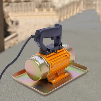 A concrete vibrator sits on a textured surface with a blurred background of a construction site.
