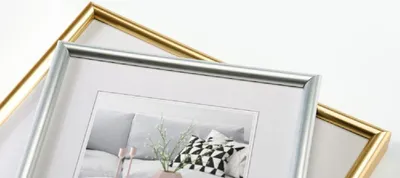 Two picture frames, one gold and one silver, are shown overlapping each other against a white background. The silver frame is in the foreground and contains a picture of a sofa with pillows.