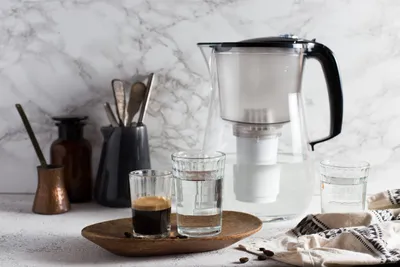 A water filter pitcher is shown on a table with glasses of water and coffee, along with some coffee-making accessories.