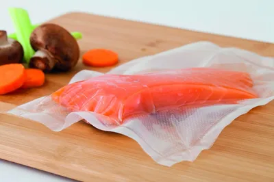 A vacuum-sealed piece of salmon is on a cutting board with some vegetables.