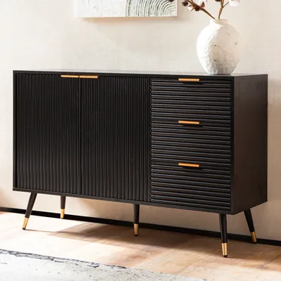 A black sideboard with gold accents and fluted detailing, standing on tapered legs with gold tips. A white vase with branches is placed on top.