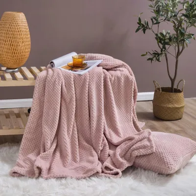 A pink blanket is draped over a wooden shelf, with a cup and saucer on top. A pillow is on the floor in front of the blanket. A plant in a basket is to the right.