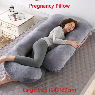 A woman is sleeping on a large U-shaped pregnancy pillow on a bed. A nightstand with a clock and a book is visible in the background.