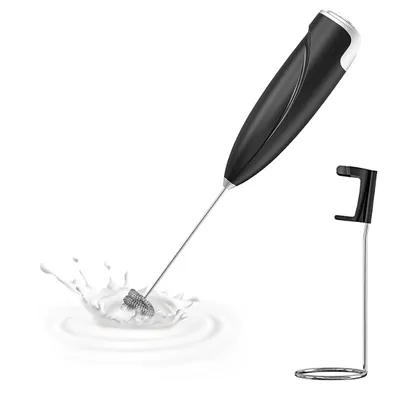 A black electric milk frother with a silver whisk attachment is shown in a splash of milk, with its black and silver stand to the right. The background is plain white.