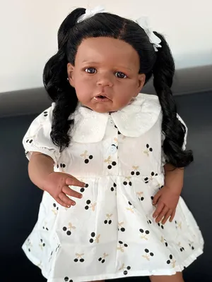 Image de Corps en coton de 60 cm, yeux bleus, longs cheveux noirs bouclés, cils collés, combinaison et chaussures blanches à motifs cerises, poupée très réaliste.
