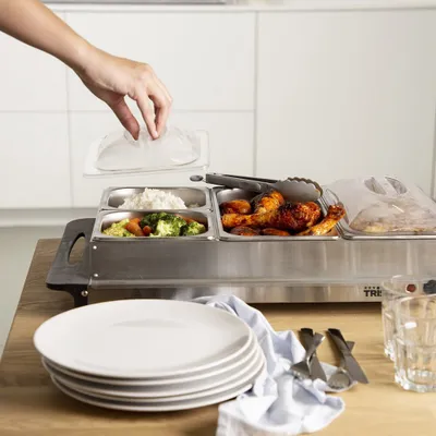 A hand is placing a lid on a buffet food warmer filled with various dishes, including rice, vegetables, and chicken.