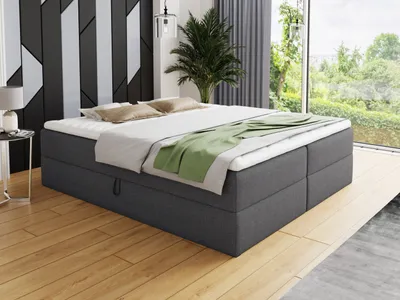 A dark gray boxspring bed with white bedding and a green throw blanket is shown in a bedroom with a modern geometric wall design and large windows overlooking a garden.