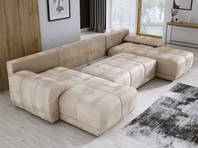 A large, U-shaped sectional sofa in a beige velvet fabric is shown in a modern living room setting. The sofa has a modular design with multiple seating sections and ottomans. A rug with a black and white abstract pattern is visible in the foreground.
