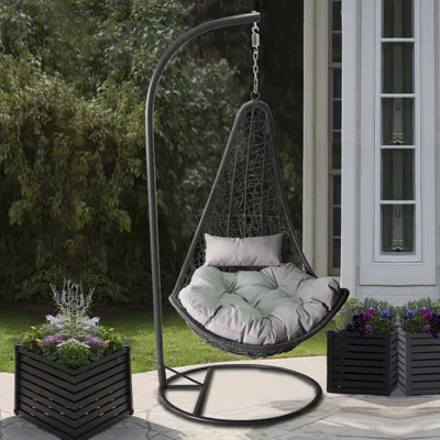 A hanging chair with cushions and pillows is placed on a patio next to planters with flowers and greenery.