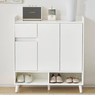 A white shoe cabinet with two doors, a drawer, and an open shelf at the bottom, displaying several pairs of shoes. A Marshall speaker and a desk calendar are on top.