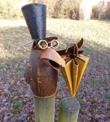 A metal sculpture of a raven wearing a top hat and glasses, holding a closed umbrella, mounted on wooden posts in an outdoor setting with fallen leaves.