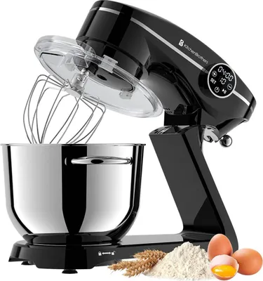 A black KitchenBrothers stand mixer with a stainless steel bowl, whisk attachment, and a transparent lid. There is also a small pile of flour and wheat stalks, and three eggs in the foreground.