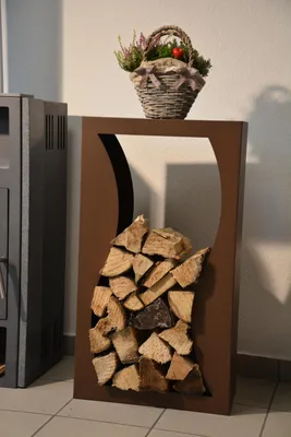 A metal firewood rack with a corten effect finish, filled with logs, stands next to a dark gray stove. A decorative basket with plants and a red ornament sits on top of the rack.