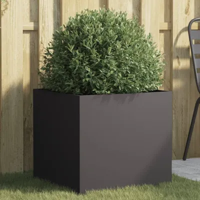 A black planter with a green bush in it, placed on grass in front of a wooden fence.