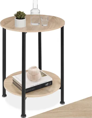 A round, two-tiered side table with a wood-look top and black metal legs, decorated with a plant, a bottle, a glass, and books.