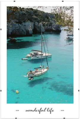 Two sailboats are anchored in a clear turquoise bay with rocky cliffs and trees in the background.