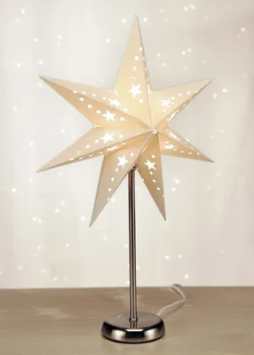 A star-shaped lamp with cut-out star patterns, illuminated from within, stands on a metallic base and stem on a wooden surface.