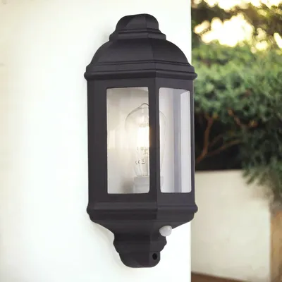 A black outdoor lantern with a motion sensor is mounted on a white wall, with greenery in the background.