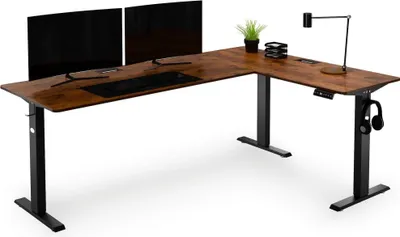 An L-shaped standing desk with two monitors, a keyboard, a lamp, and headphones.
