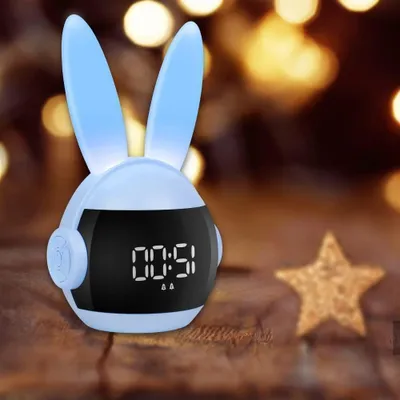 A close-up of a blue bunny-shaped alarm clock with a digital display, set on a wooden surface with blurred bokeh lights in the background and a star decoration to the right.