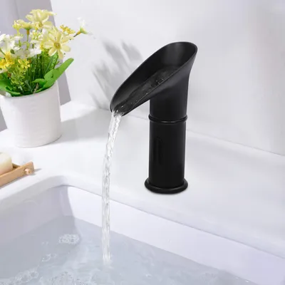 A black waterfall faucet is shown in a bathroom sink, with water flowing into the sink. A potted plant with yellow flowers is visible to the left.