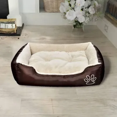 A brown and beige dog bed is placed on a light-colored wooden floor in a room with a fireplace and a vase of flowers in the background.