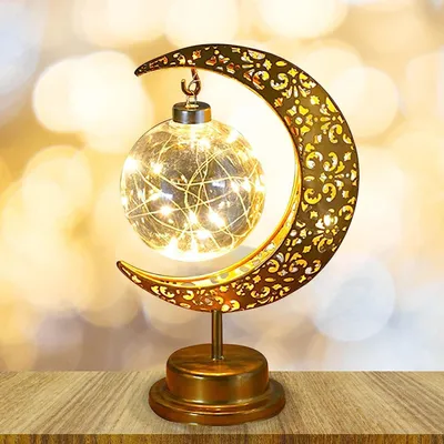 A decorative Ramadan lamp shaped like a crescent moon with a glass orb containing fairy lights, placed on a wooden surface.