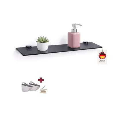 A black glass shelf mounted on a white wall, with a small potted plant and a pink soap dispenser on it. A tag with the German flag is attached to the shelf. Below the shelf, there are two shelf brackets and screws.