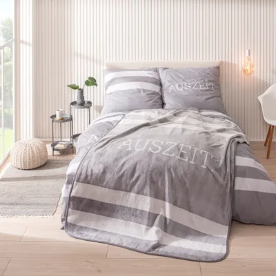 A bedroom scene with a bed made up with striped bedding and a grey fleece blanket with the word "AUSZEIT" printed on it.