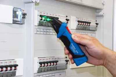 A hand is holding a voltage tester and checking a circuit breaker panel.